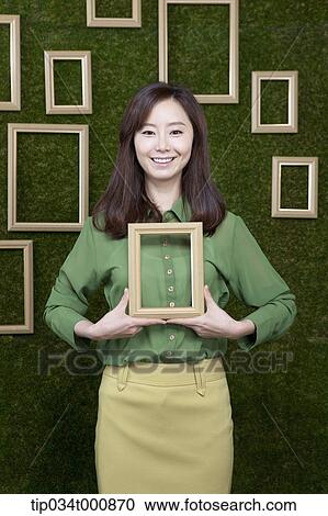 Stock Image - A woman holding a wooden frame from the green wall. Fotosearch