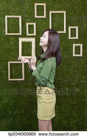 Stock Photo - A woman holding a wooden frame from the green wall. Fotosearch