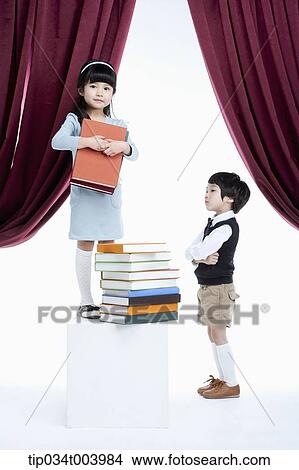 Picture - a boy standing next to a girl with pile of books on a stage. Fotosearch