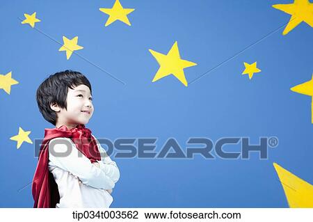 Stock Image - a boy wearing a red cape standing with arms crossed in front of a blue wall full of stars. Fotosearch