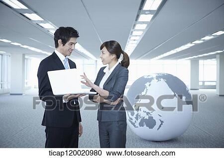 Stock Image - a businessman and woman discussing with laptop next to a huge globe in an empty room. Fotosearch