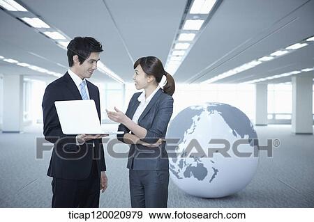 Stock Photo - a businessman and woman discussing with laptop next to a huge globe in an empty room. Fotosearch