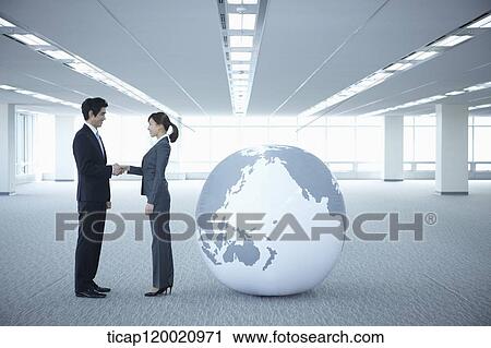 Stock Image - a businessman and woman shaking hands next to a huge globe in an empty room. Fotosearch