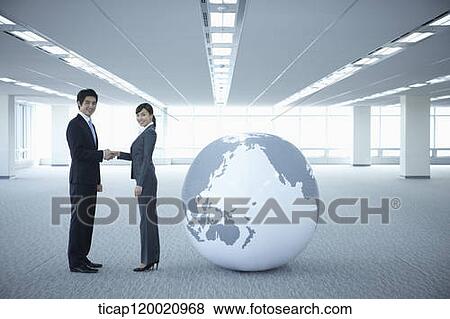 Stock Photo - a businessman and woman shaking hands next to a huge globe in an empty room. Fotosearch
