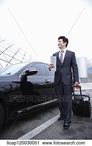 Stock Image - a businessman standing with a luggage and a passport next to a car. Fotosearch