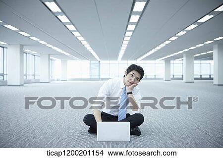Picture - a businessman using laptop on the floor of an empty room with a hand on his chin. Fotosearch