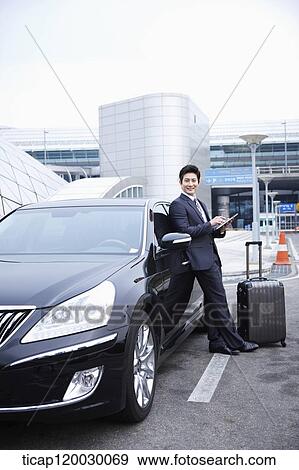 Stock Photo - a businessman with a luggage leaning on a car using a tablet PC. Fotosearch