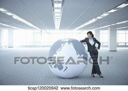 Stock Image - a businesswoman leaning on a huge globe in an empty room. Fotosearch
