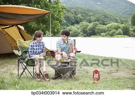 Picture - a couple sitting on camping chairs having a camping meal. Fotosearch
