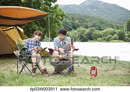 Stock Image - a couple sitting on camping chairs having a camping meal. Fotosearch