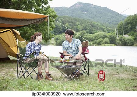 Stock Image - a couple sitting on camping chairs chatting while having a camping meal. Fotosearch