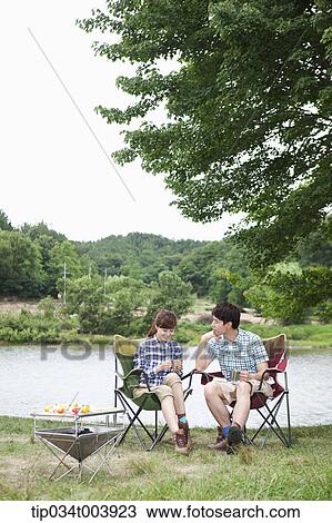 Stock Image - a couple sitting on camping chairs under a tree in front of a lake. Fotosearch