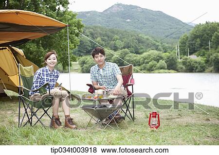 Stock Photo - a couple sitting on camping chairs smiling at camera while having a camping meal. Fotosearch