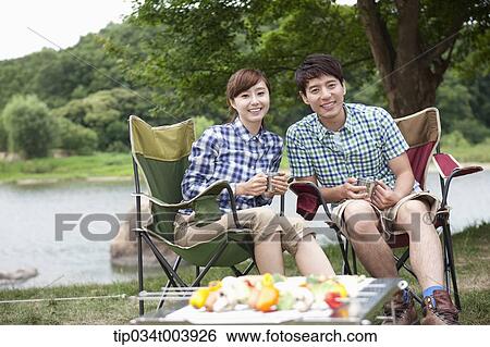 Stock Photograph - a couple sitting on camping chairs with a grill in front of them . Fotosearch