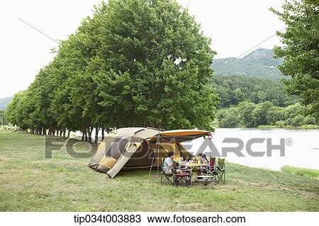 Stock Image - a family having a camping meal in camping place with trees and a lake. Fotosearch