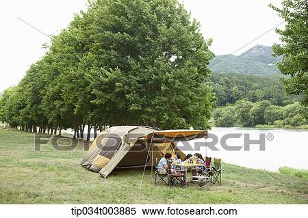 Stock Photography - a family having a camping meal in camping place with trees and a lake. Fotosearch