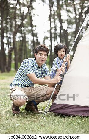 Stock Image - a father and son smiling at camera building a tent in camping place. Fotosearch