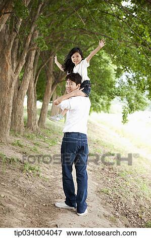 Stock Photo - a father with a daughter sitting on his shoulders walking along trees . Fotosearch