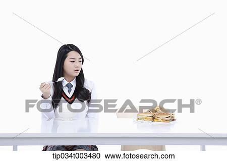Stock Image - a female student with school uniform staring at burgers while holding a spoon with rice. Fotosearch