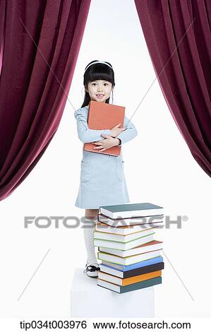 Stock Photograph - a girl holding a book while standing in front of pile of books . Fotosearch