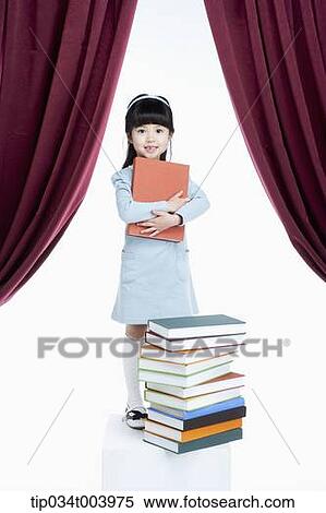 Stock Photography - a girl holding a book while standing in front of pile of books . Fotosearch