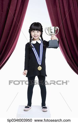 Stock Image - a girl holding a trophy cup on a stage with red curtains. Fotosearch