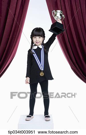 Stock Image - a girl holding a trophy cup on a stage with red curtains. Fotosearch