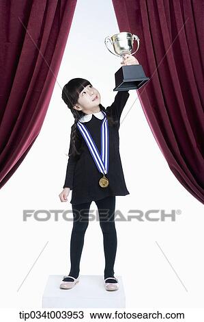 Stock Image - a girl holding a trophy cup on a stage with red curtains. Fotosearch
