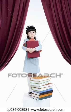 Stock Image - a girl holding books while standing in front of pile of books on a stage. Fotosearch