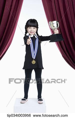 Stock Photograph - a girl with a trophy cup showing her thumbs up on a stage with red curtains. Fotosearch