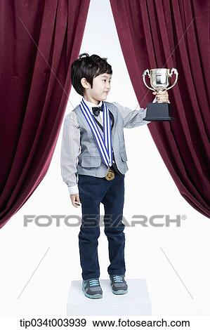 Stock Photo - a kid posing with a trophy cup in front of a red curtains. Fotosearch