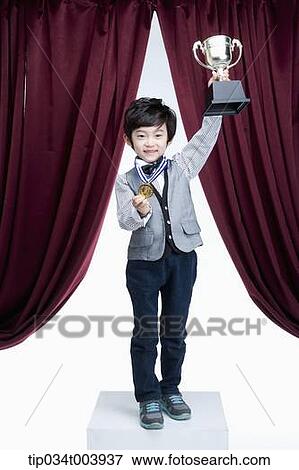 Stock Photo - a kid raising a trophy cup in front of a red curtains . Fotosearch