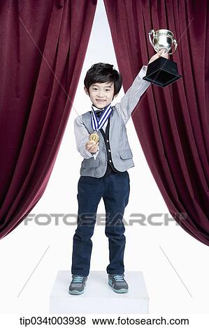 Stock Photo - a kid raising a trophy cup in front of a red curtains . Fotosearch