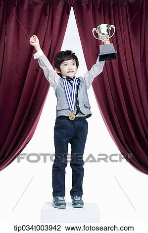 Stock Image - a kid raising hands with a trophy cup in front of a red curtains. Fotosearch
