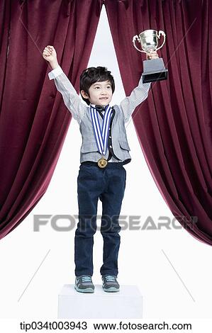 Stock Image - a kid raising hands with a trophy cup in front of a red curtains. Fotosearch