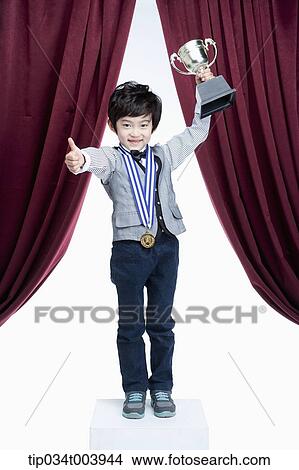Picture - a kid with trophy cup showing his thumbs up on a stage with red curtains. Fotosearch