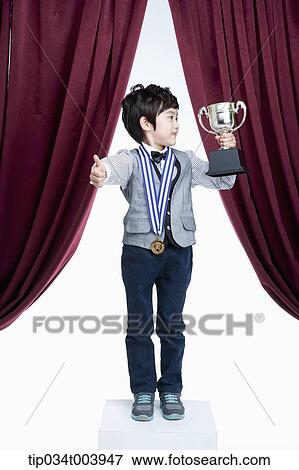 Stock Photo - a kid with trophy cup showing his thumbs up on a stage with red curtains. Fotosearch