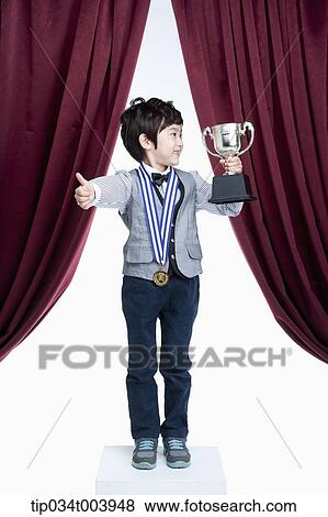 Stock Photo - a kid with trophy cup showing his thumbs up on a stage with red curtains. Fotosearch