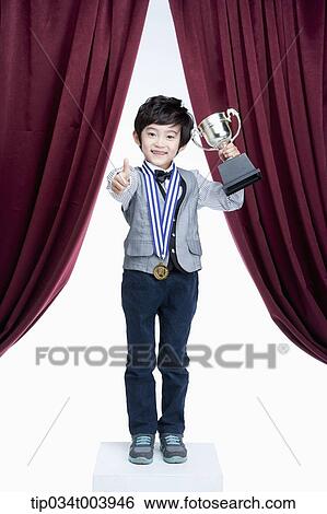 Stock Photograph - a kid with trophy cup showing his thumbs up on a stage with red curtains. Fotosearch