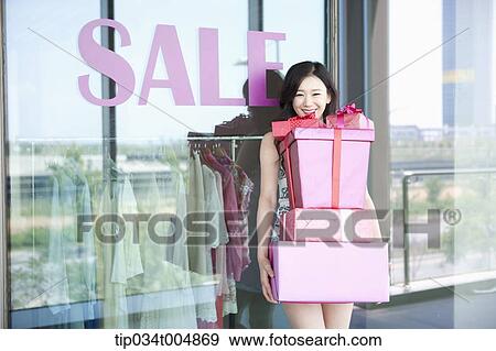 Stock Photo - a lady holding piles of presents in front of a show window with a sale sign. Fotosearch