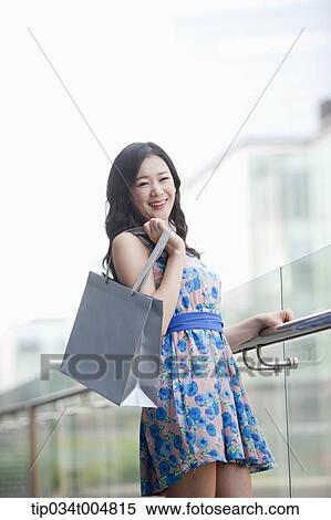 Stock Photography - a lady in flower printed dress posing while holding shopping bag. Fotosearch