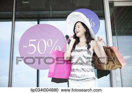 Stock Image - a lady wearing hat holding shopping bags smiling in front of a show window during sale . Fotosearch