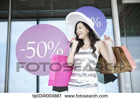 Stock Image - a lady wearing hat holding shopping bags smiling in front of a show window during sale . Fotosearch