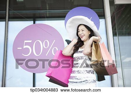 Stock Image - a lady wearing hat holding shopping bags smiling in front of a show window during sale . Fotosearch
