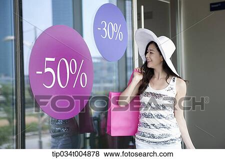 Stock Photo - a lady wearing hat holding shopping bags smiling on a show window during sale . Fotosearch