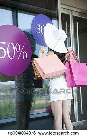 Stock Photograph - a lady wearing hat holding shopping bags looking on a show window during sale . Fotosearch