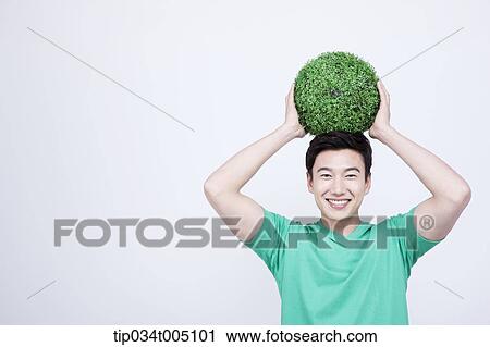 Stock Image - a man in green t-shirt holding a ball made from grass on his head. Fotosearch