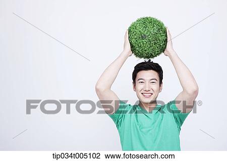 Stock Image - a man in green t-shirt holding a ball made from grass over his head. Fotosearch