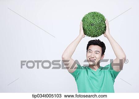 Stock Image - a man in green t-shirt holding a ball made from grass over his head. Fotosearch