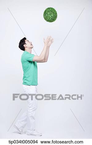 Picture - a man in green t-shirt tossing a ball made from grass . Fotosearch